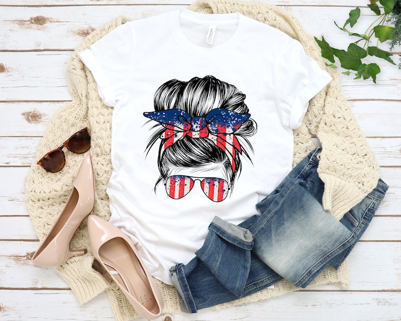 May include: White t-shirt featuring a graphic of a woman with a messy bun and a red, white and blue bandana. The bandana has a splatter paint design.