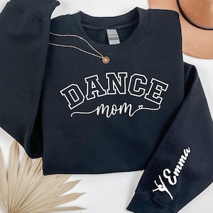 May include: Black sweatshirt with the words "DANCE mom" in white block letters. The sleeve has the name "Emma" in white script with a ballerina silhouette. A gold necklace and a tan hat are also visible.