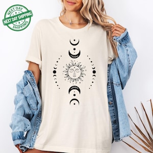 May include: Cream-colored t-shirt featuring a black sun and moon phases design. The graphic includes a smiling sun and crescent moons. The shirt is paired with a blue denim jacket. The image also includes a green and white "NEXT DAY SHIPPING" sticker.