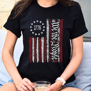 May include: A black t-shirt with a distressed American flag design and the text "We the People" and "1776".