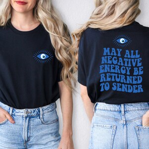 Puede incluir: Camiseta negra con un dise&ntilde;o de ojo azul en la parte delantera y trasera. La parte trasera de la camiseta presenta el texto "MAY ALL NEGATIVE ENERGY BE RETURNED TO SENDER" en azul. La camiseta es de material suave.