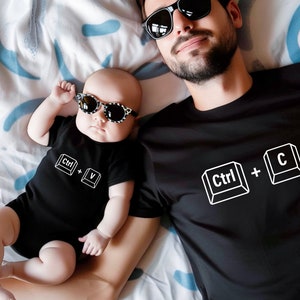 May include: A black t-shirt with the text "Ctrl + C" in white, and a black bodysuit with the text "Ctrl + V" in white. A man and a baby are wearing the shirts and looking at the camera.