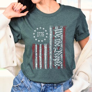 May include: A teal blue t-shirt with a distressed American flag design. The flag has red and white stripes and a circle of stars with the number 1776 in the center. The text "We the People" is written vertically on the right side of the flag.