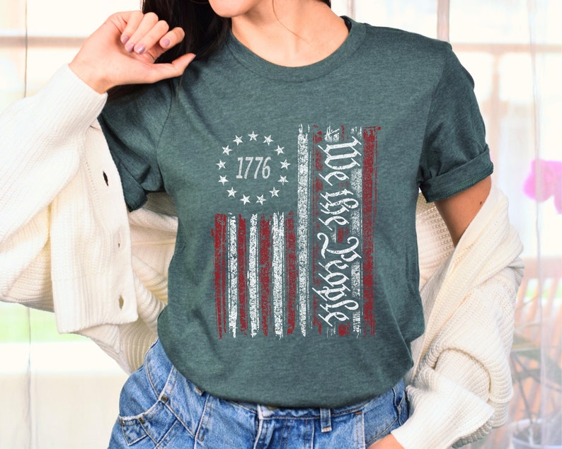 May include: A teal blue t-shirt with a distressed American flag design. The flag has red and white stripes and a circle of stars with the number 1776 in the center. The text "We the People" is written vertically on the right side of the flag.