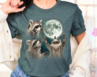 Pode incluir: Uma camiseta azul turquesa com um gráfico de três guaxinins olhando para cima para uma lua cheia. A lua é branca e os guaxinins são marrons e cinzentos.