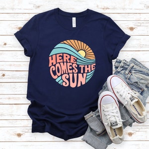 Puede incluir: Una camiseta azul marino con un gr&aacute;fico retro de un sol, olas y el texto "Here Comes The Sun".