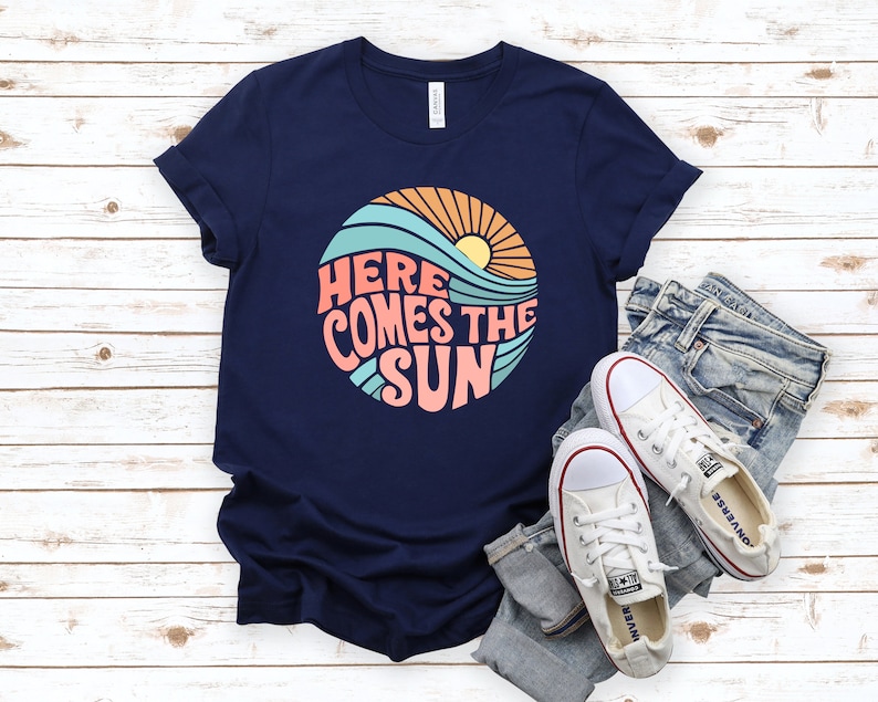 Puede incluir: Una camiseta azul marino con un gr&aacute;fico retro de un sol, olas y el texto "Here Comes The Sun".