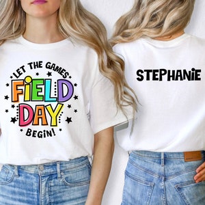 May include: White t-shirt with the text "Let the games begin! Field Day" in rainbow colors and black stars. The back of the shirt has the name "Stephanie" in black text.