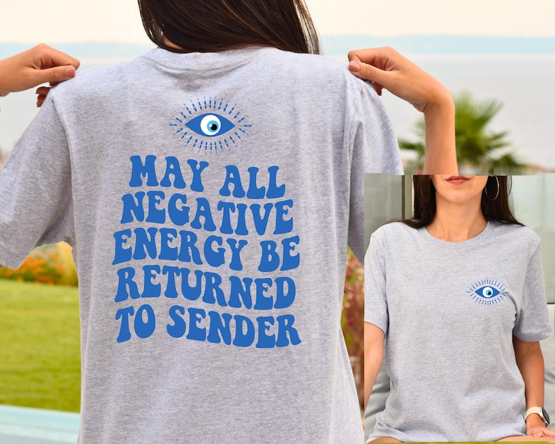 Puede incluir: Camiseta gris claro con un dise&ntilde;o de ojo azul en la parte delantera y trasera. La parte trasera de la camiseta presenta el texto "MAY ALL NEGATIVE ENERGY BE RETURNED TO SENDER" en azul, con letras de estilo retro.