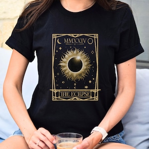 May include: Black t-shirt featuring a gold-colored solar eclipse design with the text "THE ECLIPSE" and the Roman numeral "MMXXIV". The design includes stars and a crescent moon. The shirt is made of a soft material.