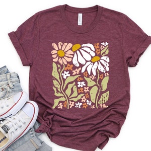 May include: A maroon t-shirt with a floral print featuring white, pink, and yellow flowers on a green stem. The flowers are stylized and have a blocky, retro look.