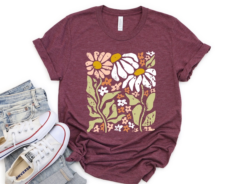 May include: A maroon t-shirt with a floral print featuring white, pink, and yellow flowers on a green stem. The flowers are stylized and have a blocky, retro look.