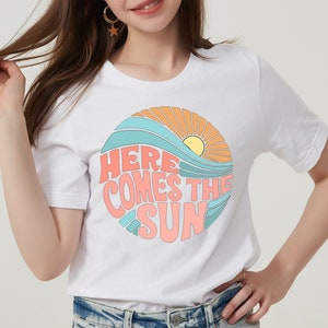 Puede incluir: Camiseta blanca con un gr&aacute;fico de un sol, olas y el texto "Here Comes The Sun" en un estilo retro.