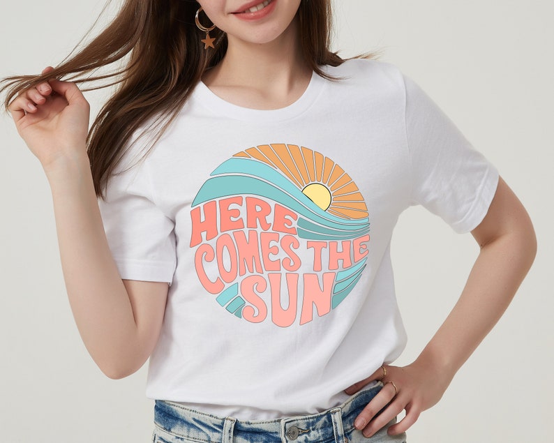 Puede incluir: Camiseta blanca con un gr&aacute;fico de un sol, olas y el texto "Here Comes The Sun" en un estilo retro.
