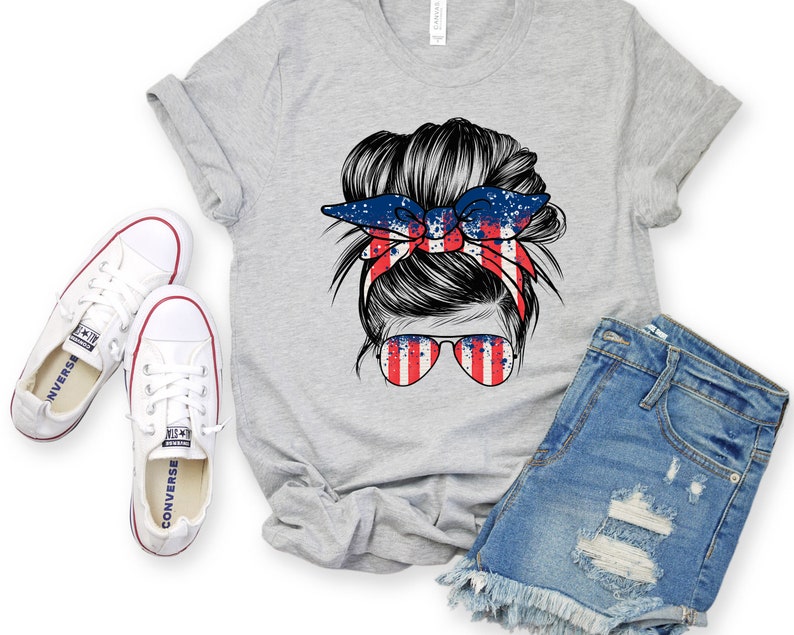 May include: A grey t-shirt featuring a graphic of a woman's hair in a messy bun with a red, white and blue bandana. The bandana has a splatter paint design. The woman is wearing sunglasses with a red, white and blue striped design.