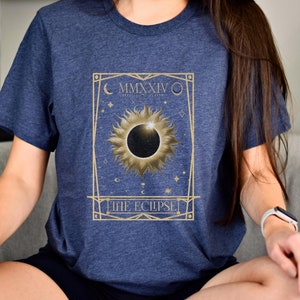 May include: Navy blue t-shirt featuring a gold-colored design of a solar eclipse with the text "THE ECLIPSE" and the Roman numeral "MMXXIV". The design includes a sun, moon, and stars.