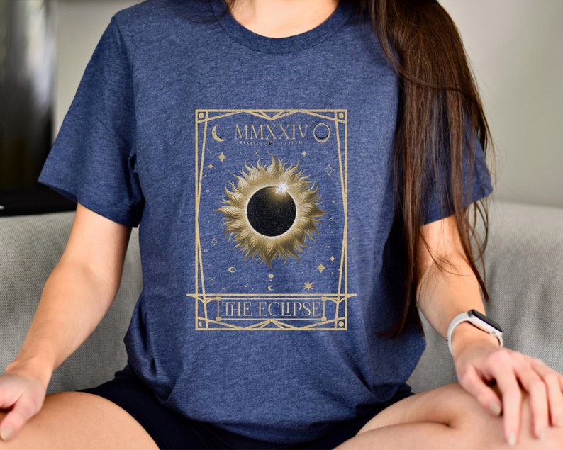 May include: Navy blue t-shirt featuring a gold-colored design of a solar eclipse with the text "THE ECLIPSE" and the Roman numeral "MMXXIV". The design includes a sun, moon, and stars.