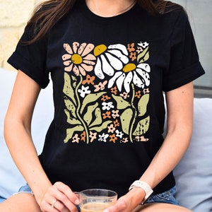 May include: A black t-shirt with a floral print featuring white, pink, and yellow daisies with green leaves and stems. The print has a distressed, vintage look.