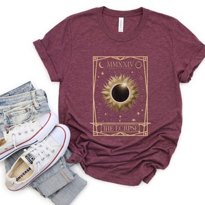 May include: Burgundy t-shirt featuring a gold-colored graphic of a solar eclipse with the text "MMXXIV" and "THE ECLIPSE". The shirt is paired with blue jeans and white sneakers.