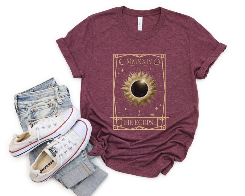 May include: Burgundy t-shirt featuring a gold-colored graphic of a solar eclipse with the text "MMXXIV" and "THE ECLIPSE". The shirt is paired with blue jeans and white sneakers.