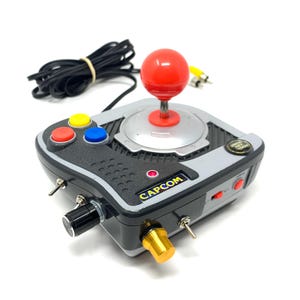 May include: A retro arcade joystick controller with a red ball-top joystick, red, yellow, and blue buttons, and the "CAPCOM" logo. The controller has a black and grey design with various switches and knobs. A black cable is coiled nearby.