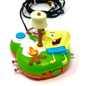 Peut inclure: Une console de jeu TV verte et orange avec SpongeBob l'Éponge, avec un chamallow sur un bâton, un feu de camp et un bouton bleu. La console présente les personnages de Bob l'éponge et Timmy Turner.