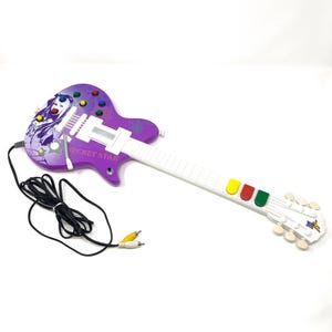 Peut inclure: Une guitare jouet violette avec un graphique représentant une personne, le texte SECRET STAR et un manche blanc doté de boutons de frettes jaune, rouge et vert. Elle comprend un câble noir avec des connecteurs RCA jaune et blanc.