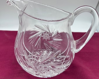 May include: A clear glass pitcher with a cut glass design. The pitcher has a round body and a handle.