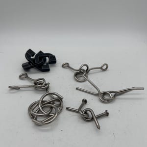 May include: A collection of metal brain teaser puzzles in various shapes. The puzzles include interlocking rings, bent wires, and a black chain-like puzzle. The objects are displayed against a white background.