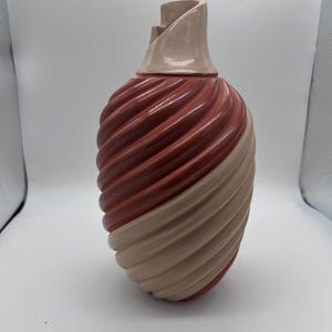 Puede incluir: Un jarrón de cerámica con un diseño en espiral. El jarrón es una combinación de marrón rojizo y beige, con un patrón texturizado y acanalado. El cuello del jarrón es beige y tiene una forma angular única.