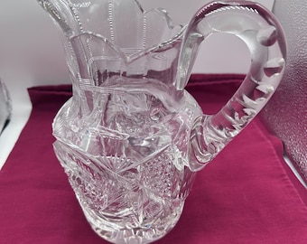 May include: A clear glass pitcher with a cut glass design. The pitcher has a wide mouth and a handle.