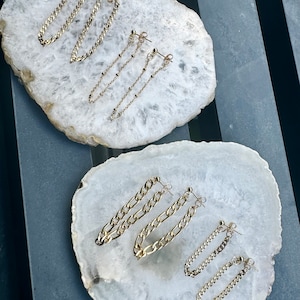 May include: Gold-toned chain earrings in various styles, including curb chain and beaded designs. The earrings are displayed on a white, marbled surface, showcasing their different lengths and textures. The earrings are presented in pairs.