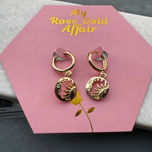 May include: Gold-coloured hoop earrings with a sun and moon pendant. The earrings are displayed on a pink hexagonal card with the text "My Rose Gold Affair" in gold lettering. The earrings are on a dark surface.