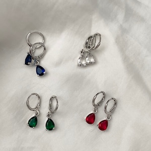 May include: Four pairs of silver hoop earrings with dangling teardrop-shaped gemstones. The earrings feature blue, clear, green, and red gemstones.