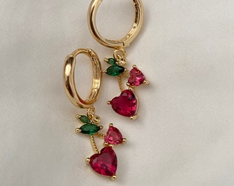 May include: Gold hoop earrings with a dangling charm featuring red and green heart-shaped crystals. The charm resembles a cherry.