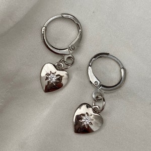 Puede incluir: Un par de pendientes de aro de plata con un pequeño dije de corazón colgando de cada aro. Cada dije de corazón tiene un pequeño strass transparente en el centro.