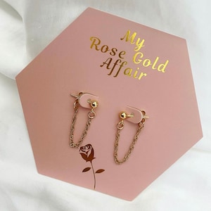 May include: A pair of gold chain earrings with small gold balls on a pink display card with the text "My Rose Gold Affair".