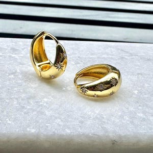 May include: A pair of gold hoop earrings with small, clear stones set in the shape of stars. The earrings are slightly curved and have a polished finish.