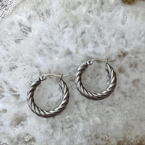 May include: A pair of silver-colored hoop earrings with a twisted rope design. The earrings are circular and have a simple clasp closure. The earrings are displayed on a white and gray surface.