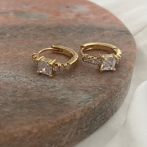 May include: A pair of gold-coloured hoop earrings. Each earring has a square, clear gemstone at the centre, surrounded by smaller, clear gemstones. The earrings are displayed on a pink and brown surface.