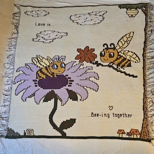 Puede incluir: Manta de ganchillo con un diseño de abeja y flor. La manta incluye el texto "Love is... Bee-ing together".
