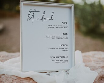 May include: A white sign with black text that reads "Let's Drink" and lists different types of alcoholic and non-alcoholic beverages. The sign is sitting on a white cloth and a brown rock.