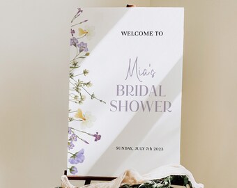 May include: White sign with the text "Welcome to Mia's Bridal Shower" and the date "Sunday, July 7th 2023". The sign is decorated with purple and yellow flowers.