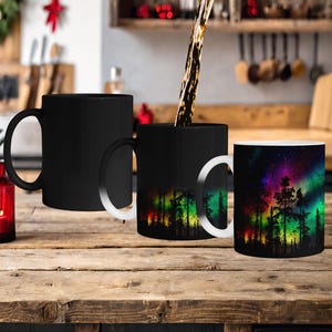 Può includere: Tre tazze nere con un designo che cambia colore. Le tazze sono su un tavolo di legno. Una tazza è piena di un liquido scuro, rivelando un'immagine colorata di un bosco con un cielo stellato e aurore boreali. Le altre due tazze sono vuote, mostrando l'esterno nero.