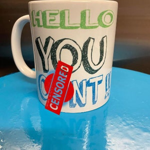 May include: White ceramic mug with a green, black and red design. The mug says "Hello You Can't" with a red sticker that says "CENSORED" over the word "Can't".