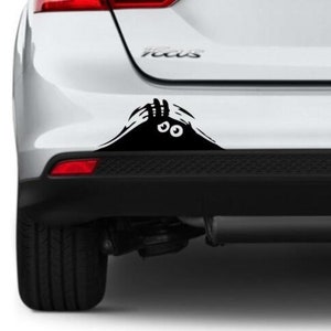 Puede incluir: Pegatina de coche en blanco y negro de un par de ojos que miran desde detrás de una forma negra. La pegatina está colocada en el parachoques trasero de un coche blanco.
