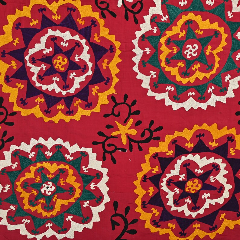 K&ouml;nnte beinhalten: Roter Stoff mit einem sich wiederholenden Muster aus bunten gestickten Blumen. Die Blumen haben eine Sternform in der Mitte und sind von konzentrischen Kreisen in Gelb, Wei&szlig;, Gr&uuml;n und Lila umgeben.