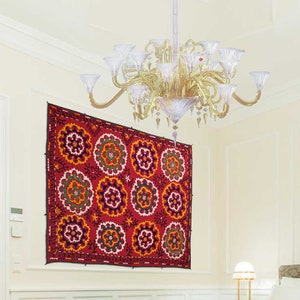 K&ouml;nnte beinhalten: Ein roter und goldener Wandteppich mit einem sich wiederholenden Muster aus bunten Kreisen h&auml;ngt an einer wei&szlig;en Wand. Ein Kristall-Kronleuchter mit goldenen Akzenten h&auml;ngt von der Decke &uuml;ber dem Wandteppich.