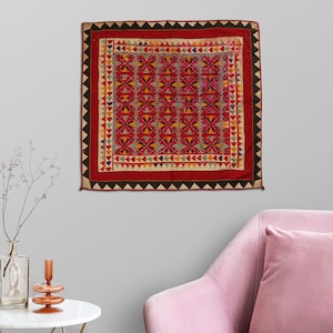 May include: A red, black, and white embroidered tapestry with geometric patterns. The tapestry is framed with a black border and has a fringe around the edges.