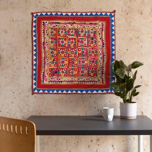 May include: A colorful, hand-stitched textile wall hanging with a geometric pattern in shades of red, yellow, green, and blue. The textile is framed by a blue and white border.